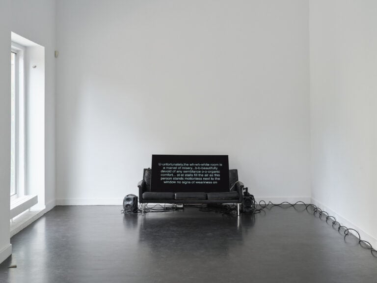 Foto: eine schwarze Ledercouch steht vor einer weißen Wand. Ein Monitor mit einem Text ist auf die Sitzfläche gestellt. Rechts und links stehen Lautsprecher. Kabel führen rechts aus dem Bild heraus