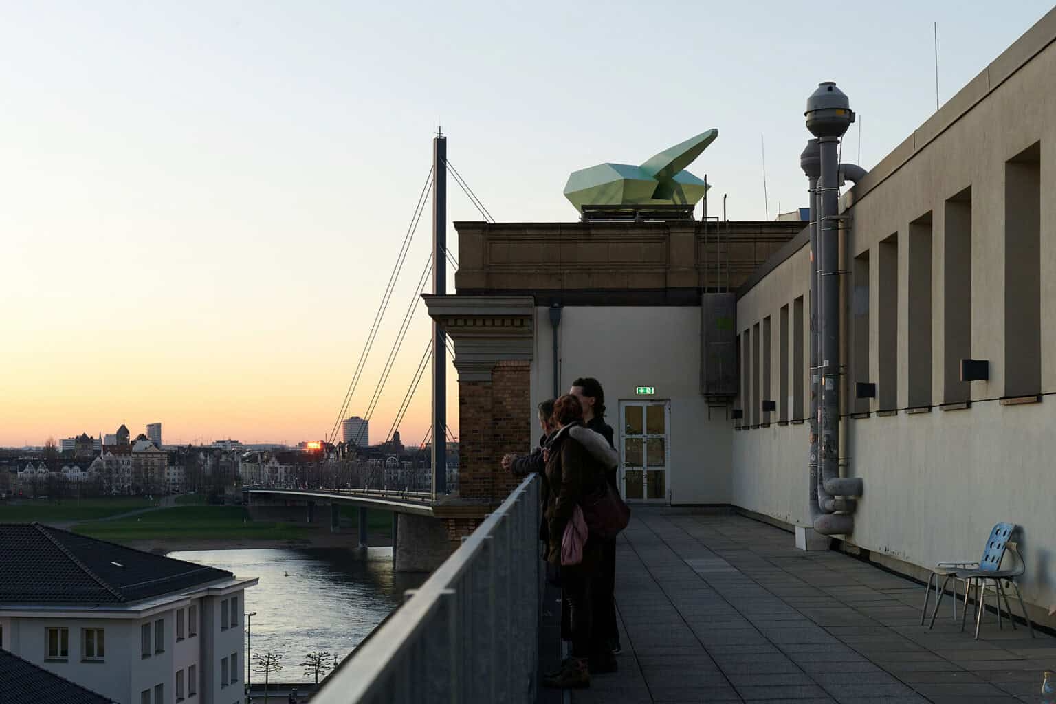 Foto: Ansicht einer Terrasse mit Ausblick auf eine Landschaft. Auf dem Hausdach steht ein Objekt. Auf der Terrasse befinden sich Menschen und Stühle.