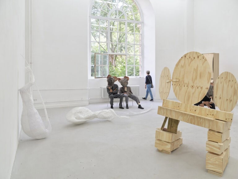 Foto: Ausstellungsraum mit mehreren Kunstwerken: Stühle mit menschgroßen Figuren mit Mausköpfen, Holzskulptur und Kunststoffskulpturen