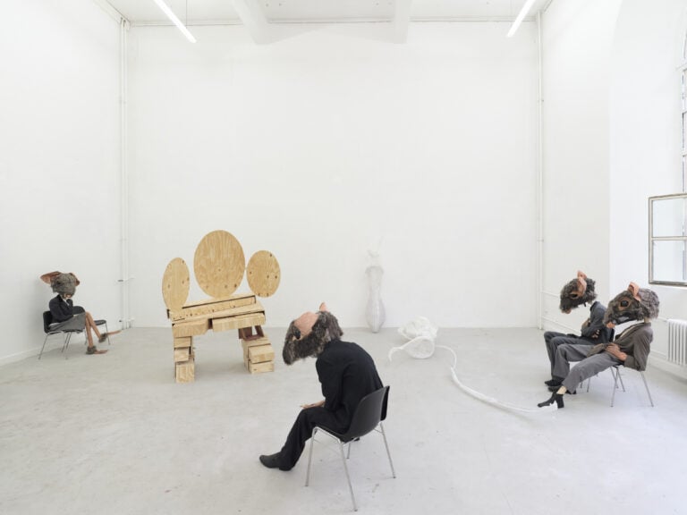 Foto: Ausstellungsraum mit mehreren Kunstwerken: Stühle mit menschgroßen Figuren mit Mausköpfen, Holzskulptur und Kunststoffskulpturen