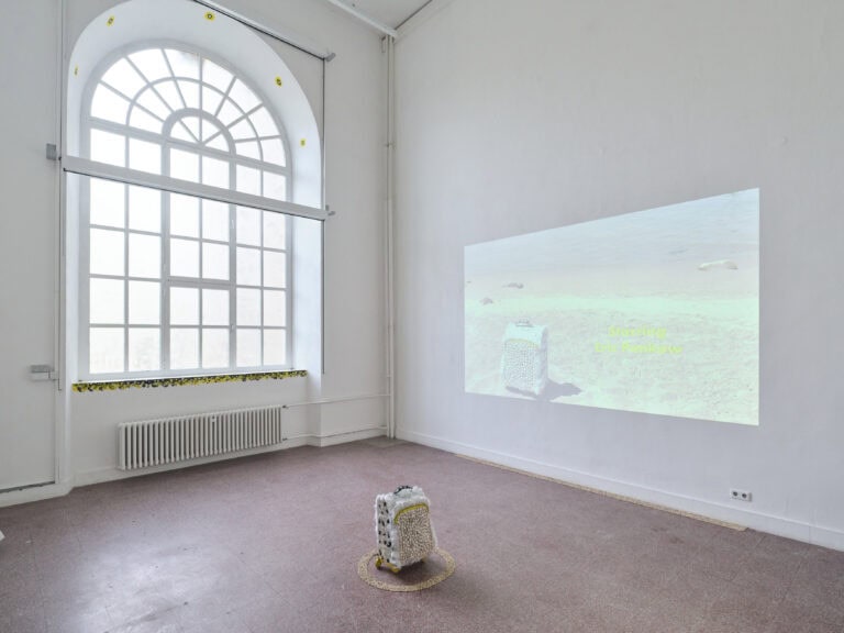 Foto: Ansicht eines Ausstellungsraums mit Fliesenboden, weißen Wänden und einem großen Fenster. In der Mitte steht ein Rollkoffer mit Federn, Samen, Muscheln beklebt in einem Ring aus weiteren Samen. An die Wand ist die Projektions eines Films zu sehen. Die Momentaufnahme zeigt den Koffer aus dem Raum.