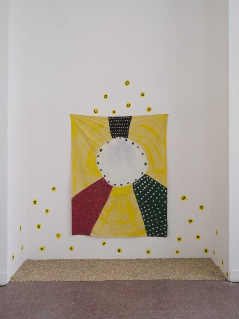 Foto: Abstrakte textile Wandarbeit an einer weißen Wand. Im Zentrum befindet sich ein weißer Kreis, umgeben von gelben, roten, grünen und schwarzen Stoffflächen. Kleine gelbe Kreise sind rund um das Werk auf der Wand verteilt.