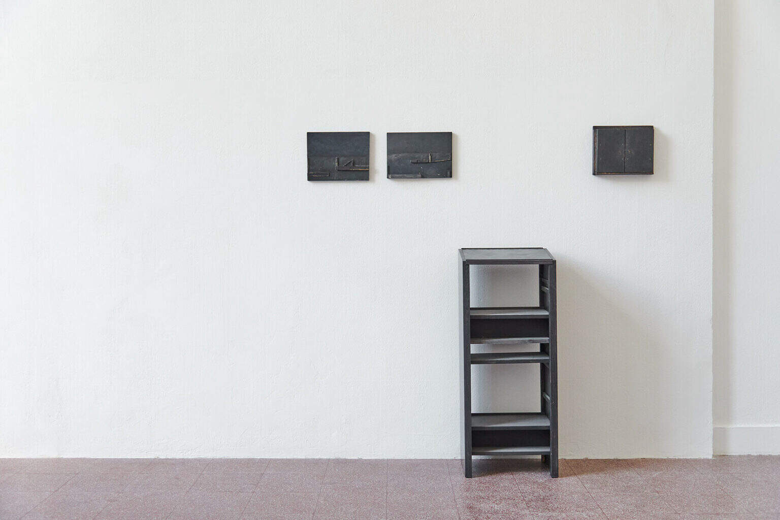 Foto: Ein Raum mit weißen Wänden und rotem Steinboden. An den Wänden des Raumes hängen, auf ähnlicher höhe, kleine, dunkle Objekte. Vor einem der Objekte auf dem Boden steht ein dunkles, pultartiges Objekt auf dem Boden.