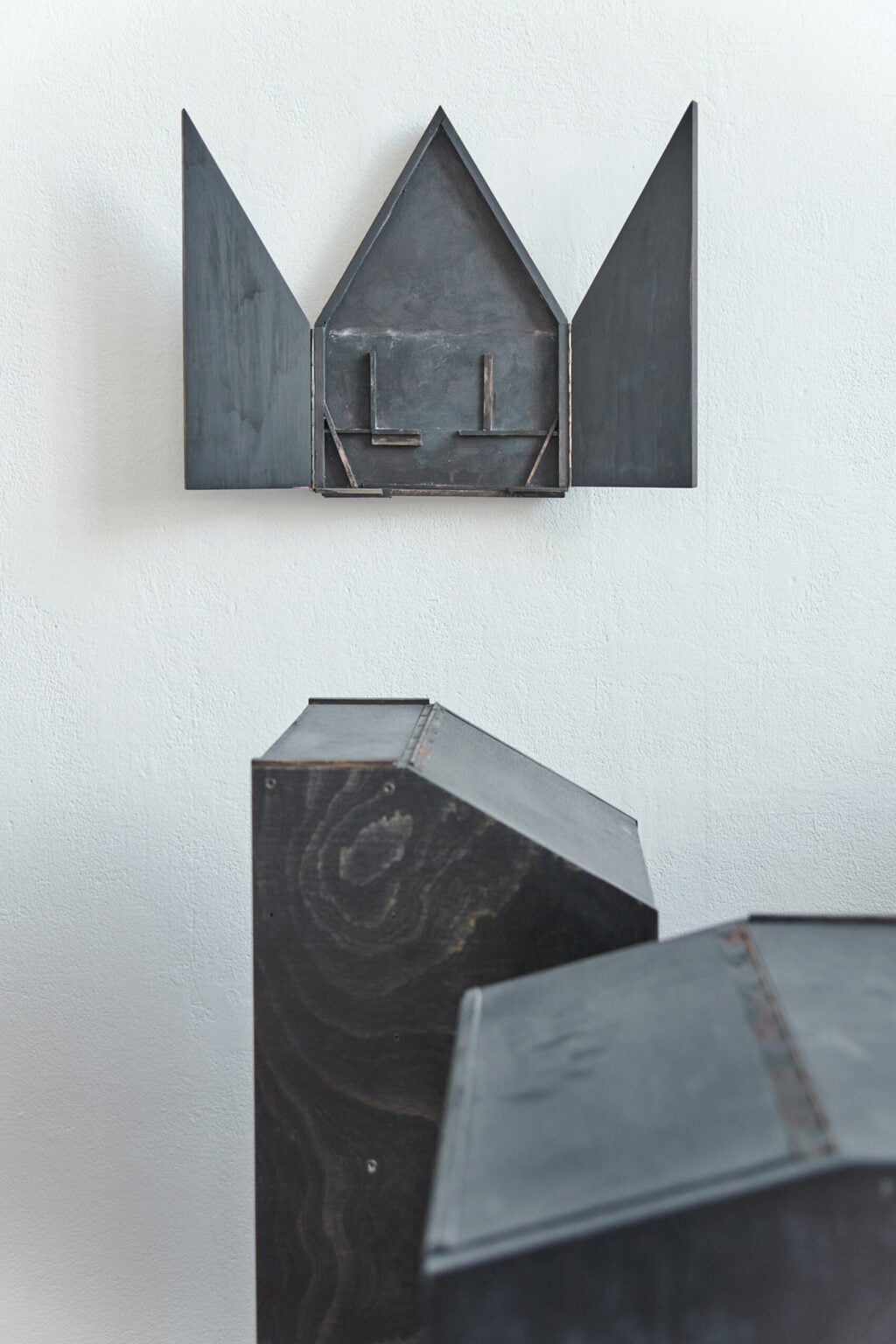 Foto: Ein Raum mit weißen Wänden und rotem Steinboden. An der Wände des Raumes hängt ein dunkles Objekt. Vor dem Objekt auf dem Boden steht eine Gruppe dunkler, pultartiger Objekte, die nah beieinander stehen.