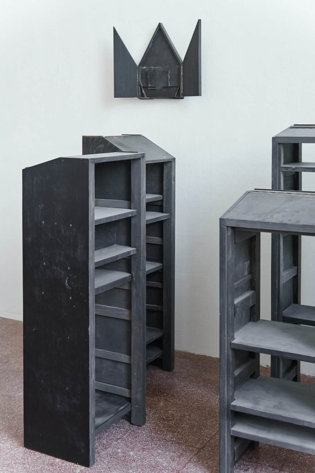 Foto: Ein Raum mit weißen Wänden und rotem Steinboden. An der Wände des Raumes hängt ein dunkles Objekt. Vor dem Objekt auf dem Boden steht eine Gruppe dunkler, pultartiger Objekte, die nah beieinander stehen.