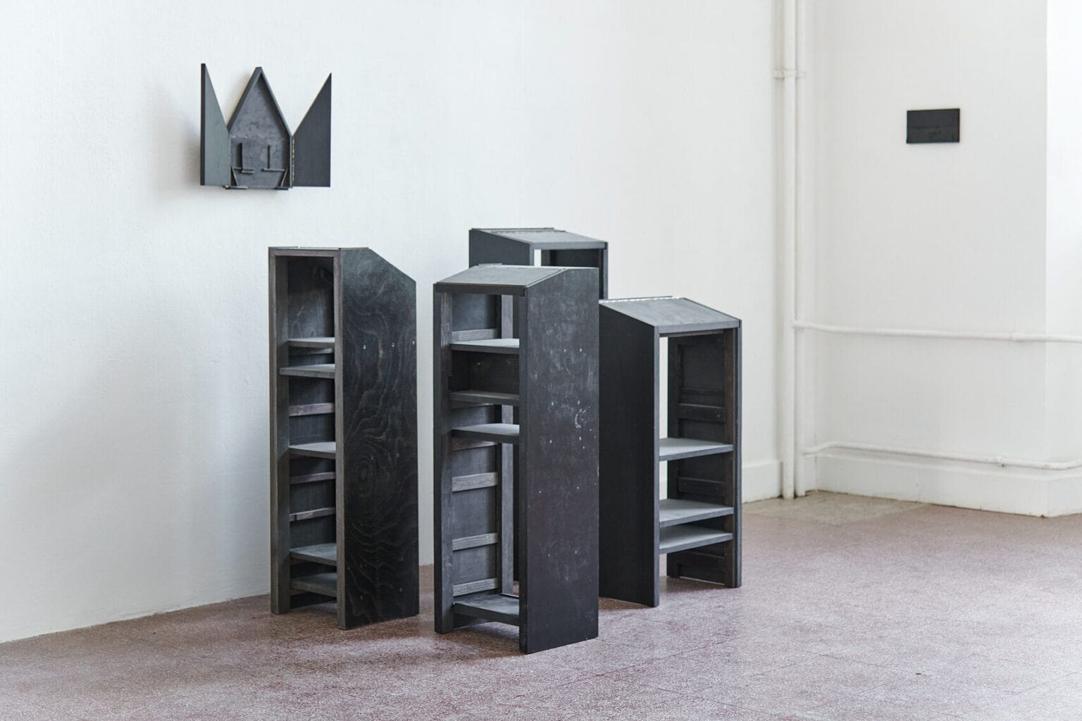 Foto: Ein Raum mit weißen Wänden und rotem Steinboden. An den Wänden des Raumes hängen, auf ähnlicher höhe, kleine, dunkle Objekte. Vor einem der Objekte auf dem Boden steht eine Gruppe dunkler, pultartiger Objekte, die nah beieinander stehen.