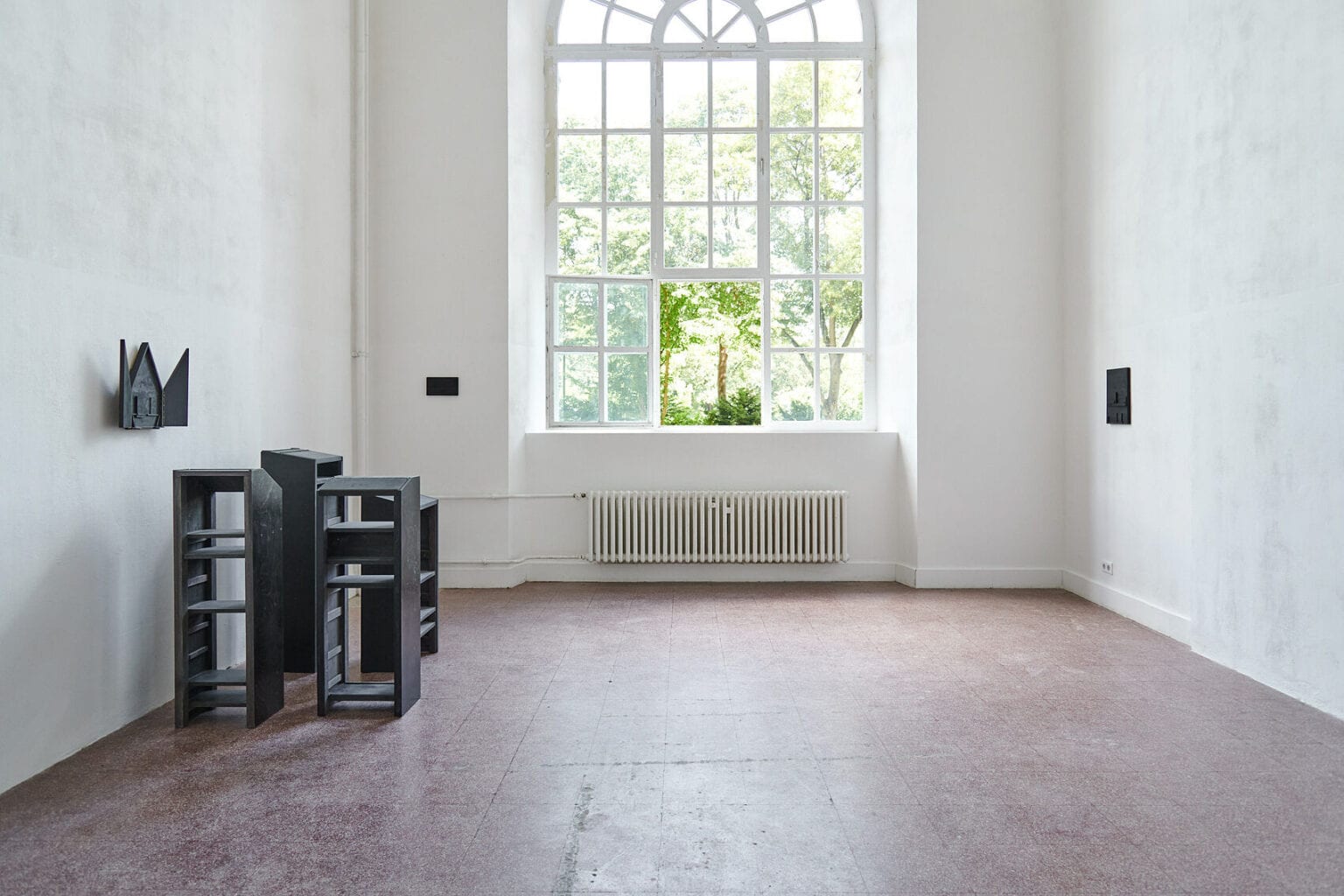 Foto: Ein Raum mit weißen Wänden, rotem Steinboden und einem großen Fenster. An den Wänden des Raumes hängen, auf ähnlicher höhe, kleine, dunkle Objekte. Vor einem der Objekte auf dem Boden steht eine Gruppe dunkler, pultartiger Objekte, die nah beieinander stehen.
