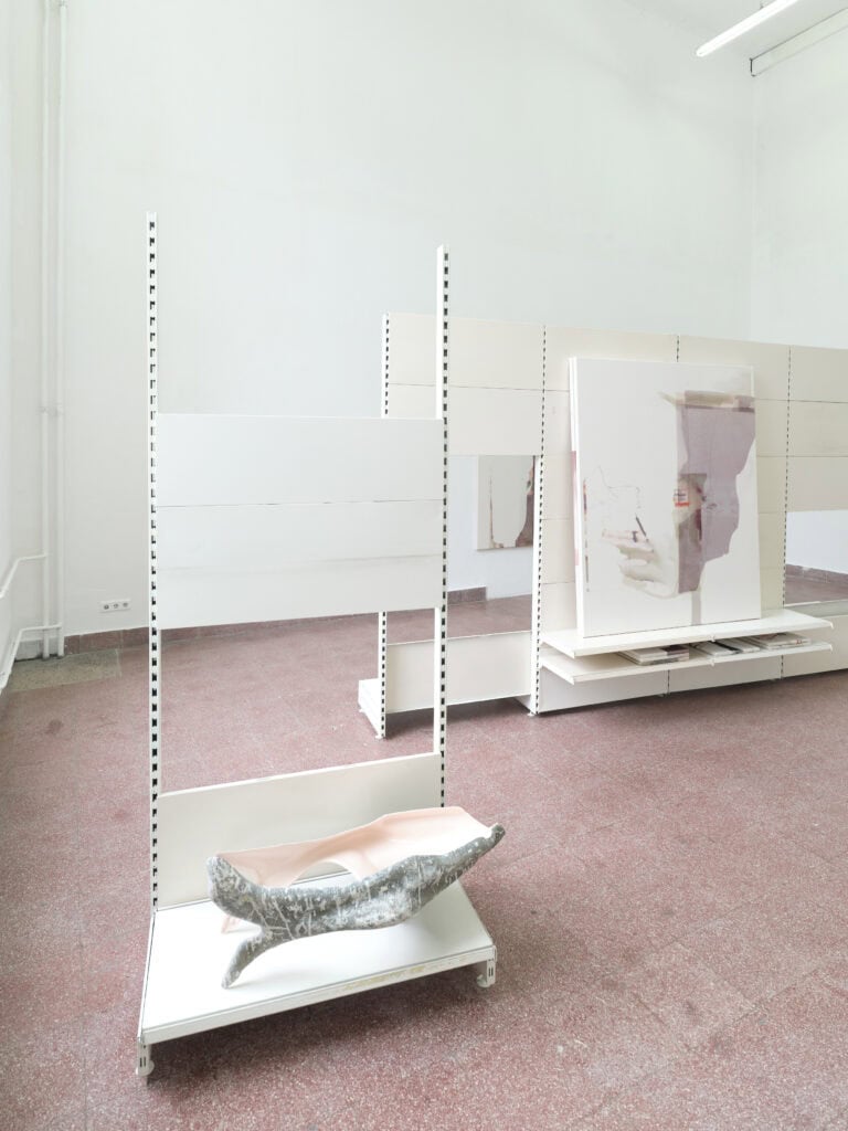 Foto: In einem hellen Ausstellungsraum mit Fliesenboden stehen weiße, bewegliche Stellwände, an denen großformatige Gemälde präsentiert werden. Weitere Kunstwerke hängen an den Wänden, und ein skulpturales Objekts liegt auf einem Regalelement.