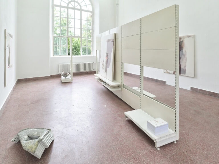 Foto: Heller Ausstellungsraum mit großem Rundbogenfenster. Im Raum stehen weiße, bewegliche Stellwände, an denen großformatige Gemälde präsentiert werden. Weitere Kunstwerke hängen an den Wänden, und zwei skulpturale Objekte liegen beziehungsweise stehen auf dem Boden.