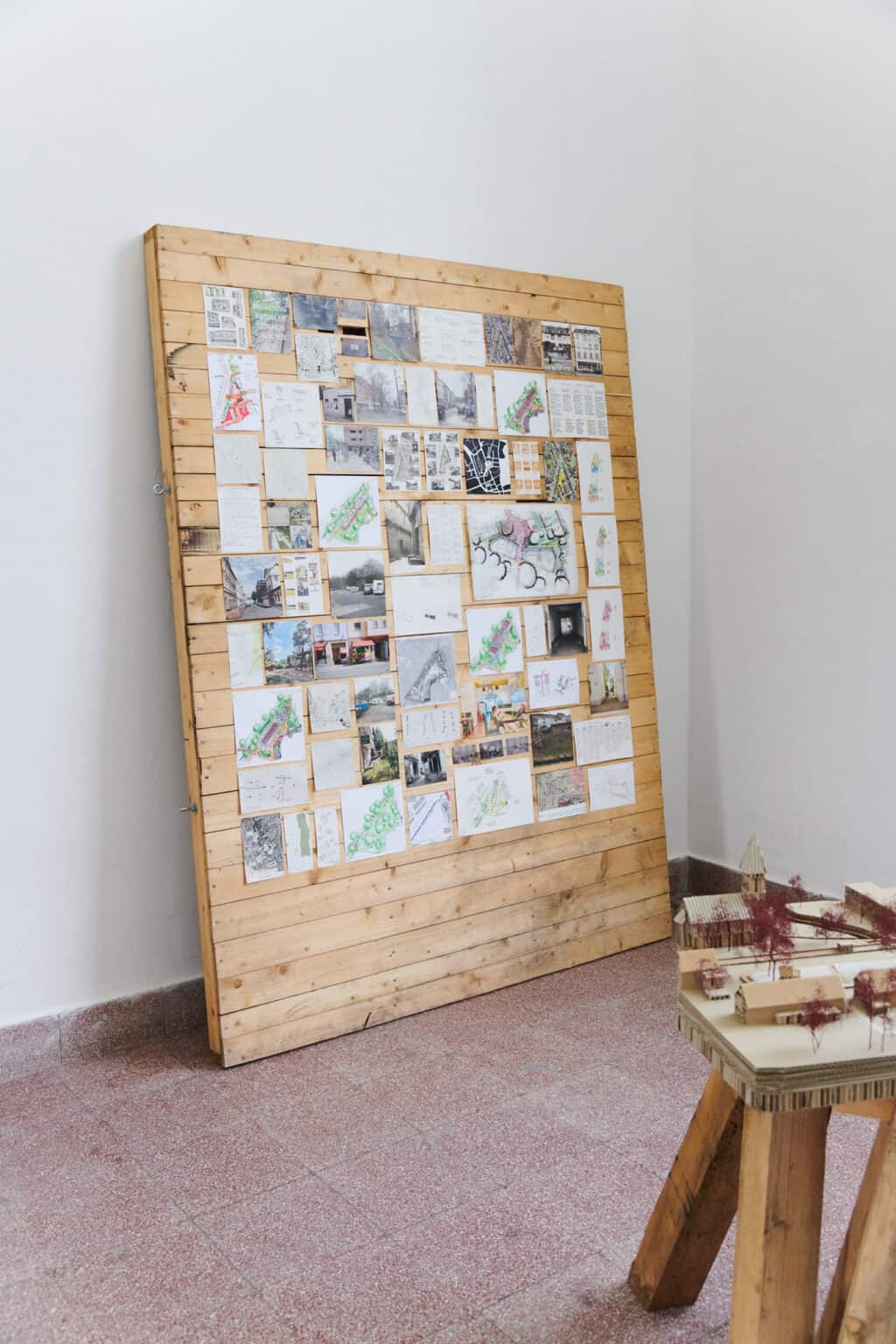 Foto: Eine Tafel aus Holzlatten, die an der Wand lehnt. Auf der Konstruktion sind viele bemalte, kleinformatige Papiere angebracht. Im Raum eine Skulptur.