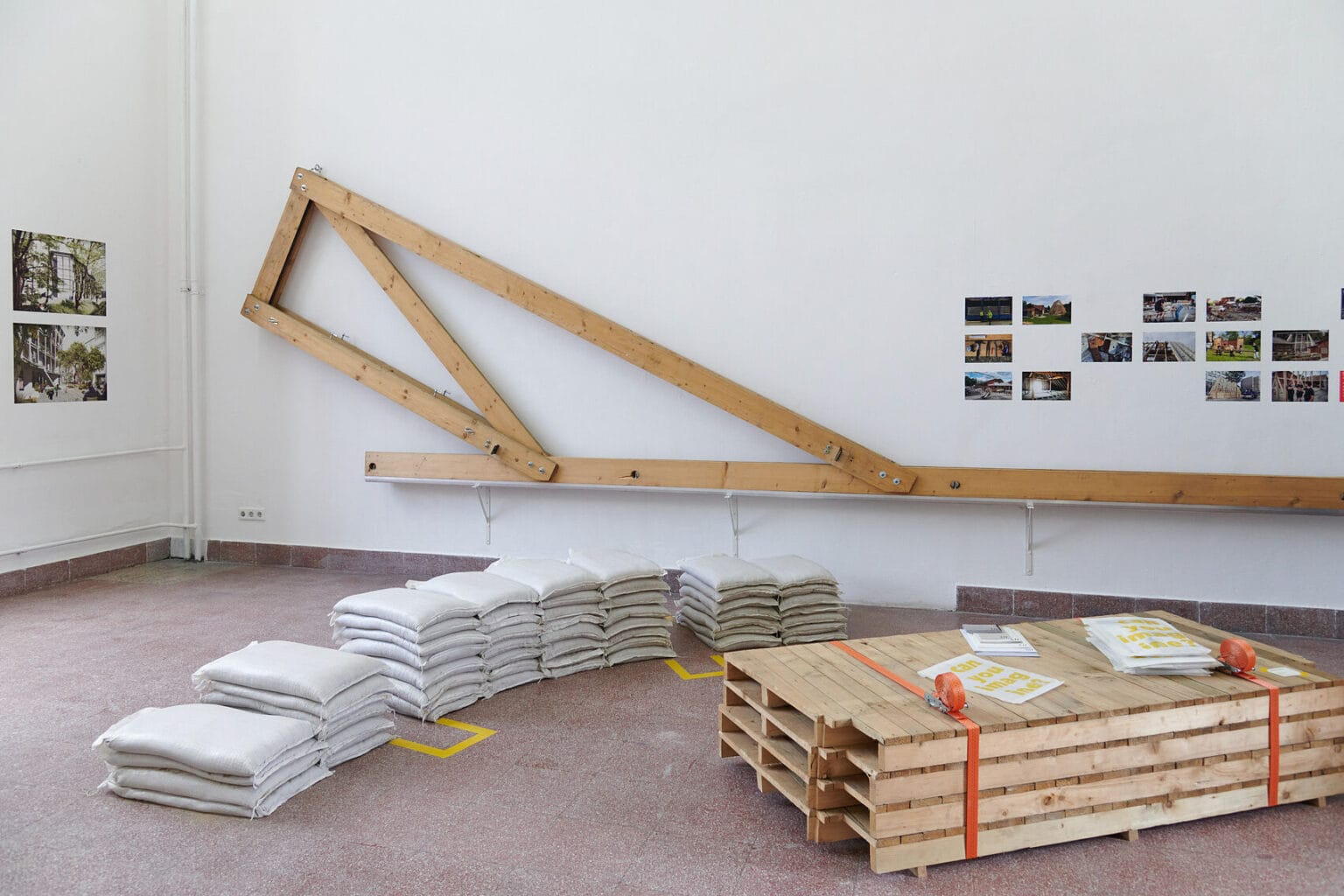 Foto: Ein Raum mit weißen Wänden und rotem Steinboden. An den Wänden des Raumes befindet sich eine große Konstruktion aus Holzlatten und Bilder in Klein- und Mittelformaten. Auf dem Boden des Raumes liegen Stapel aus gefüllten Säcken und zusammengeschnürte, aufeinander gestapelte Paletten. Auf den Paletten liegen Objekte.