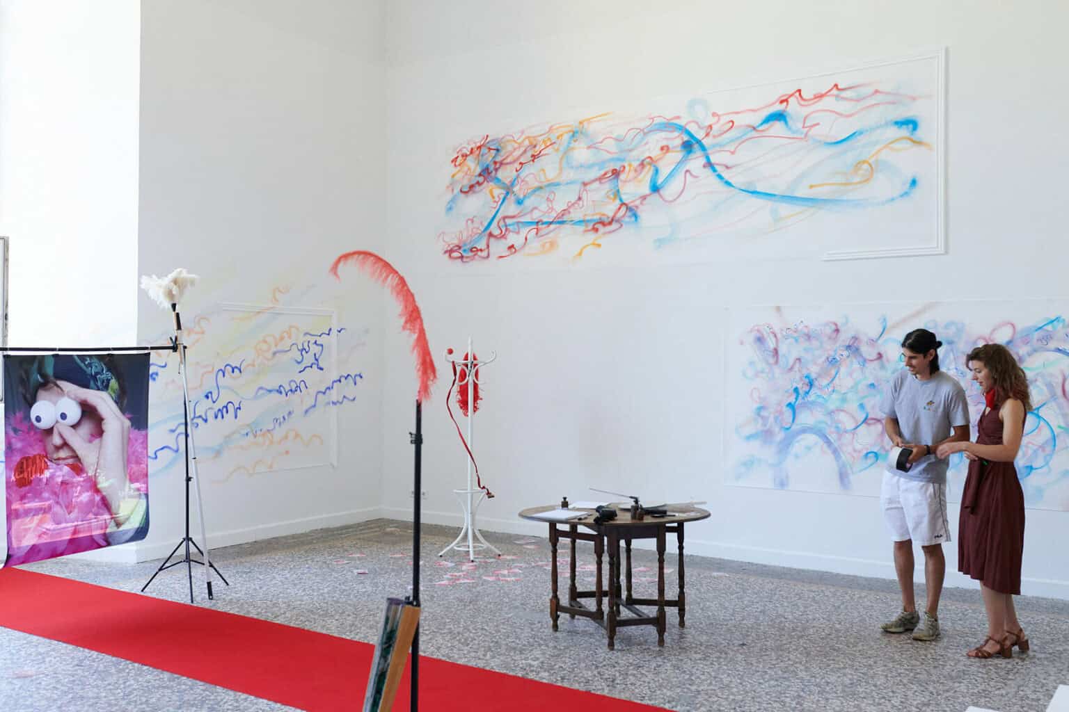 Foto: Ein Raum mit weißen Wänden, mehrfarbigem Steinboden und einem großen Fenste. Teile der Wände sind mit bunten Linien bemalt. Im Raum verteilt befindet sich eine Vielzahl unterschiedlicher Objekte, darunter ein ausgerollter, roter Teppich, Lautsprecher, ein Ventilator, ein Tisch, eine Kleiderstange, eingepackte Leinwände, Pappboxen usw. Durch den Raum bewegen sich zwei Menschen, die mit den Objekten interagieren.