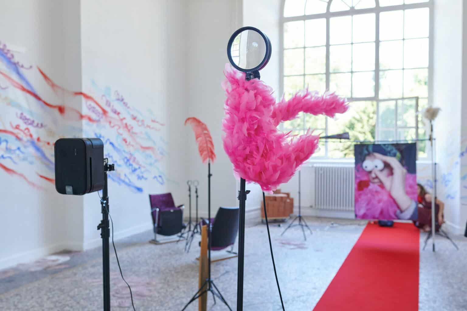 Foto: Ein Raum mit weißen Wänden, mehrfarbigem Steinboden und einem großen Fenste. Teile der Wände sind mit bunten Linien bemalt. Im Raum verteilt befindet sich eine Vielzahl unterschiedlicher Objekte, darunter ein ausgerollter, roter Teppich, Lautsprecher, ein Ventilator, ein Tisch, eine Kleiderstange, eingepackte Leinwände, Pappboxen usw.