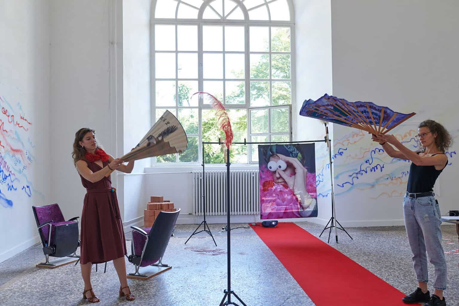 Foto: Ein Raum mit weißen Wänden, mehrfarbigem Steinboden und einem großen Fenste. Teile der Wände sind mit bunten Linien bemalt. Im Raum verteilt befindet sich eine Vielzahl unterschiedlicher Objekte, darunter ein ausgerollter, roter Teppich, Lautsprecher, ein Ventilator, ein Tisch, eine Kleiderstange, eingepackte Leinwände, Pappboxen usw. Durch den Raum bewegen sich zwei Menschen, die mit den Objekten interagieren.