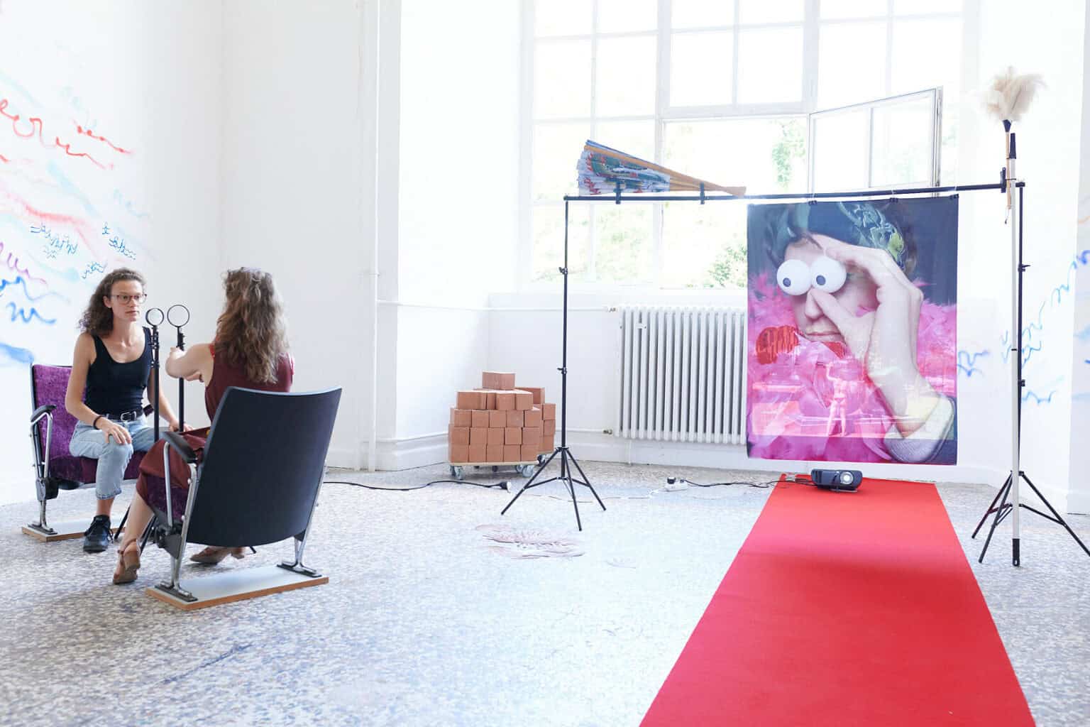 Foto: Ein Raum mit weißen Wänden, mehrfarbigem Steinboden und einem großen Fenste. Teile der Wände sind mit bunten Linien bemalt. Im Raum verteilt befindet sich eine Vielzahl unterschiedlicher Objekte, darunter ein ausgerollter, roter Teppich, Lautsprecher, ein Ventilator, ein Tisch, eine Kleiderstange, eingepackte Leinwände, Pappboxen usw. Durch den Raum bewegen sich zwei Menschen, die mit den Objekten interagieren.