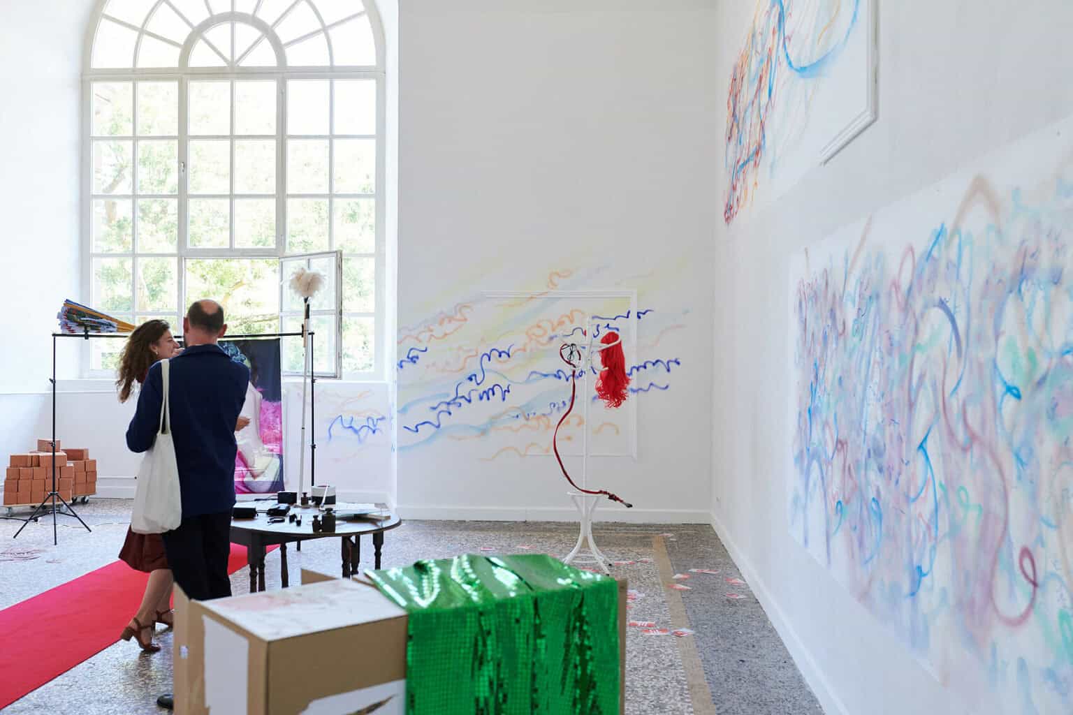 Foto: Ein Raum mit weißen Wänden, mehrfarbigem Steinboden und einem großen Fenste. Teile der Wände sind mit bunten Linien bemalt. An einer der Wände hängen flache, ebenfalls bemalte Objekte. Im Raum verteilt befindet sich eine Vielzahl unterschiedlicher Objekte, darunter ein ausgerollter, roter Teppich, Lautsprecher, ein Ventilator, ein Tisch, eine Kleiderstange, eingepackte Leinwände, Pappboxen usw. Durch den Raum bewegen sich zwei Menschen, die mit den Objekten interagieren.