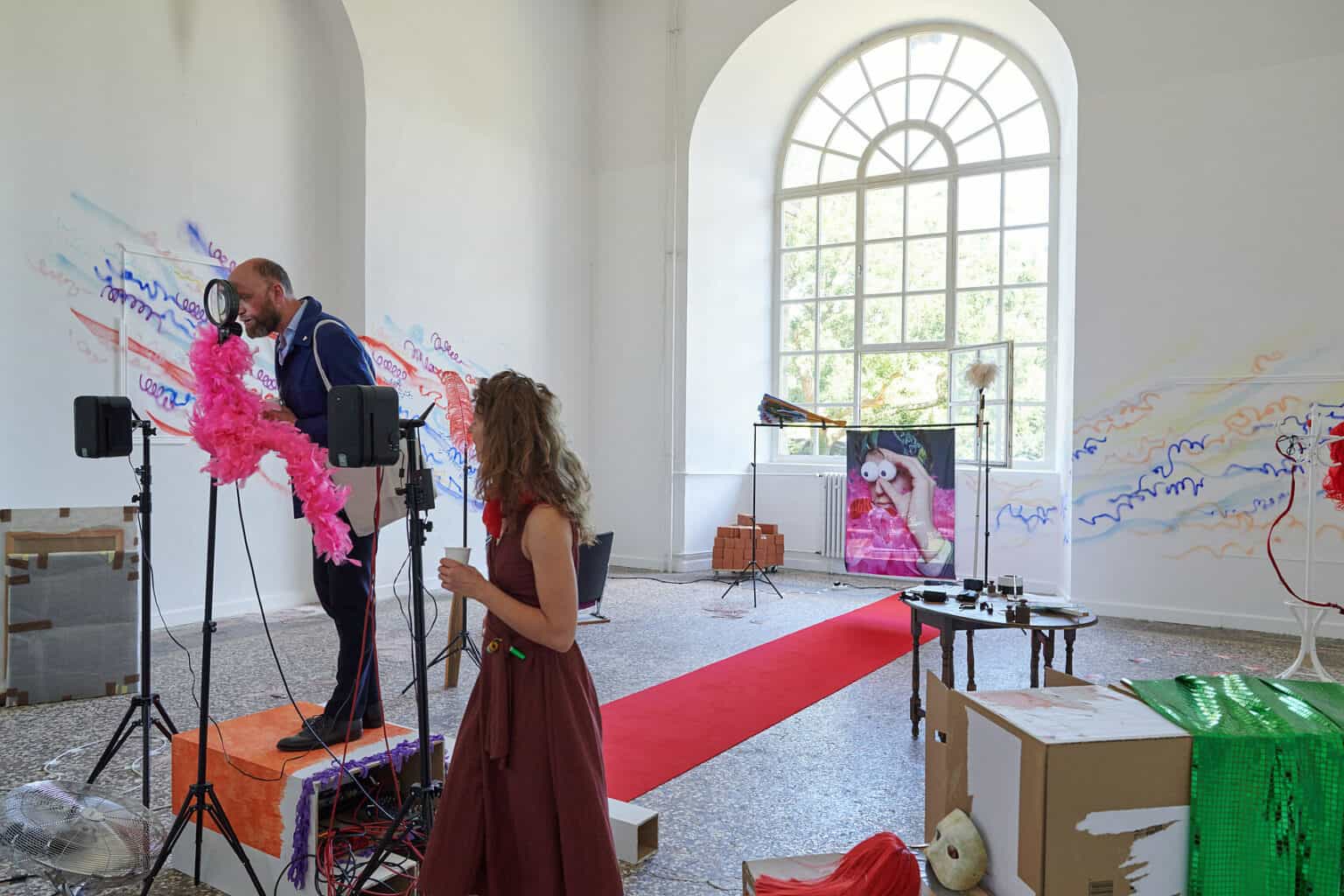 Foto: Ein Raum mit weißen Wänden, mehrfarbigem Steinboden und einem großen Fenste. Teile der Wände sind mit bunten Linien bemalt. Im Raum verteilt befindet sich eine Vielzahl unterschiedlicher Objekte, darunter ein ausgerollter, roter Teppich, Lautsprecher, ein Ventilator, ein Tisch, eine Kleiderstange, eingepackte Leinwände, Pappboxen usw. Durch den Raum bewegen sich zwei Menschen, die mit den Objekten interagieren.