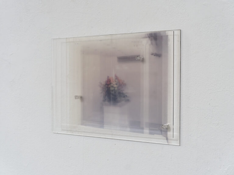 Foto: Nahaufnahme von einem Bild an einer weißen Wand. Das Bild besteht aus Glasscheiben und ein Blumenarrangement ist abgebildet.