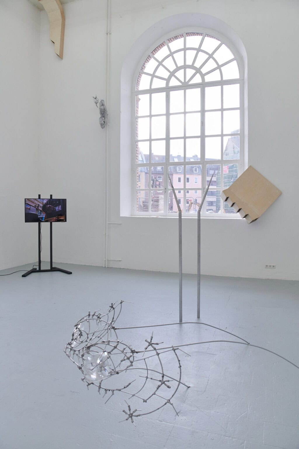 Foto: Ansicht eines Raume mit weißen Wänden, grauem Boden und großem Bogenfenster. An der Wand auf unterschiedlicher Höhe und auf dem Boden befindet sich Objekte aus unterschiedlichen Materialien.