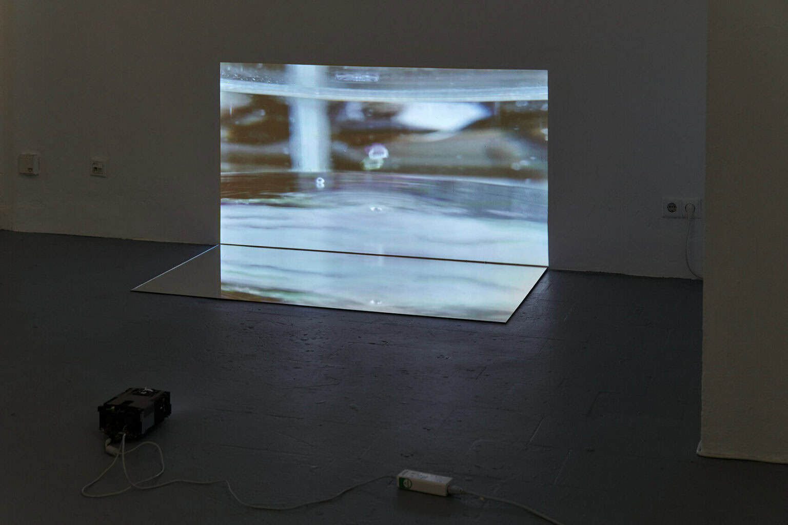Foto: Ein abgedunkelter Raum. Auf dem Boden steht ein Beamer, der bodennah ein Bild an die Wand wirft. Vor der Projektion liegt ein Spiegel auf dem Boden, der das an die Wand geworfene Bild reflektiert.