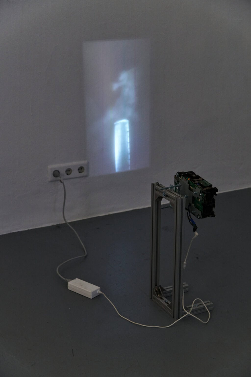 Foto: Ein abgedunkelter Raum. Im Raum steht wandnah ein kleines Gerüst, an dem ein Beamer angebracht ist. Der Beamer wirft eine Projektion an die Wand.