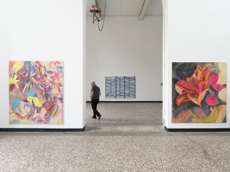 Foto: Aufnahme eines Ausstellungsraums mit Blick in den nächsten Raum durch einen großen Durchgang. Rechts und links sind an den Wänden des Durchgangs großformatige bunte Gemälde zu sehen.