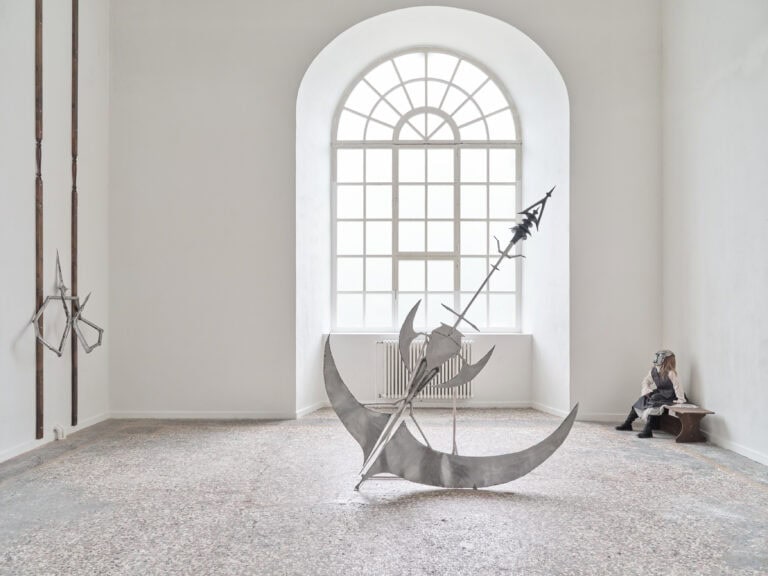 Foto: Heller Ausstellungsraum mit großem Rundbogenfenster. Im Raum stehen bzw. hängen mehrere Kunstwerke auf dem Boden und an den Wänden. Mittig ist eine große metallene Skulptur aufgestellt, die an eine Waffe erinnert. Im hinteren rechten Bereich steht eine Bank mit einer Schaufensterpuppe, die eine Metallmaske trägt. Eine spitze Metallskultptur hängt an der linken Wand.