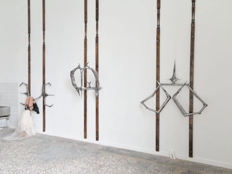 Foto: Detailaufnahme einer Wandinstallation. Sechs lange Holzstreben sind paarweise angebracht. Unterschiedliche spitze Metallelemente sind daran befestigt. Eine Schaufensterpuppe hängt kopfüber in dem linken Metallelement.