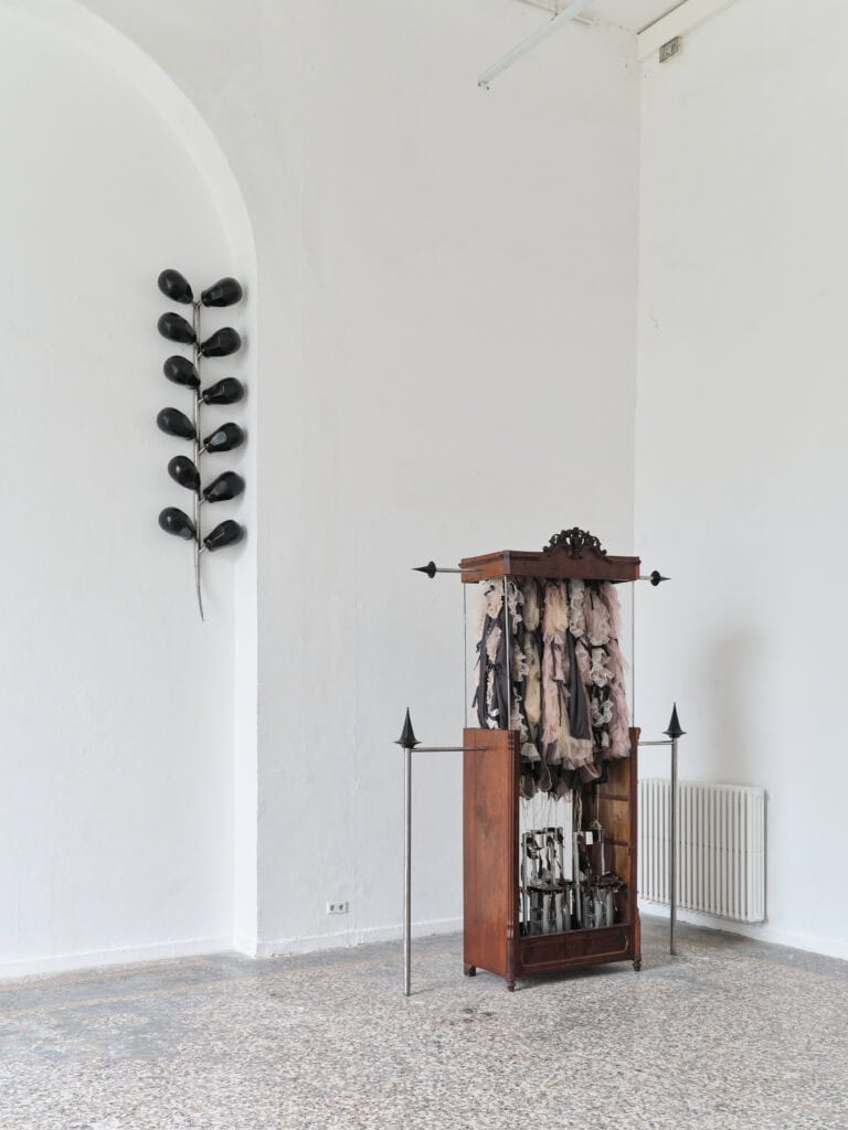Foto: Heller Ausstellungsraum mit einer Installation. Schrankartiges Modell mit Metallelementen, Kleidungsstücken. Eine weitere Metallskulptur mit schwarzen kugelförmigen Bauteilen.