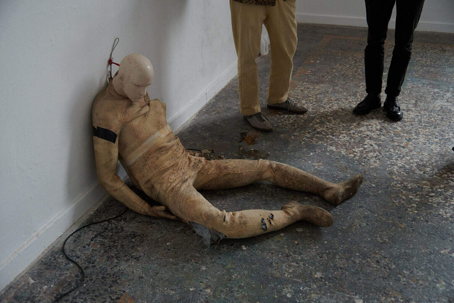 Foto: Detailaufnahme eines beschädigten und verschmutzen Crashtest-Dummys, dessen Rückenteil an einer Wand lehnt. Daneben die Beine zweier Menschen.