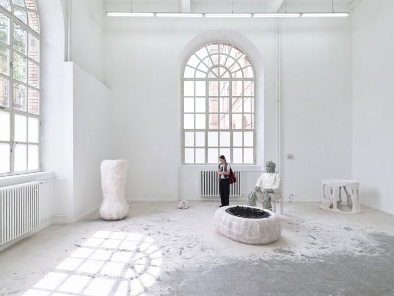 Foto: Heller Ausstellungsraum mit zwei großen Rundbogenfenstern. Im Raum stehen mehrere Kunstwerke auf dem Boden. Der Boden ist mit Sand ausgestreut. Eine Person betrachtet die Kunstwerke.