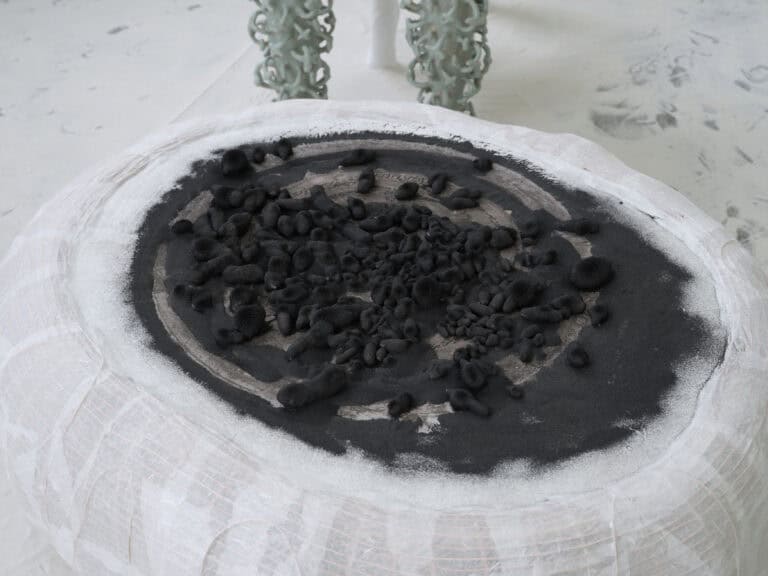 Foto: Nahaufnahme eines weißen runden Kunstwerks, auf dessen Oberseite sich diverse organische schwarze kleinere Formen aus einem sandartigen Material gebildet haben.