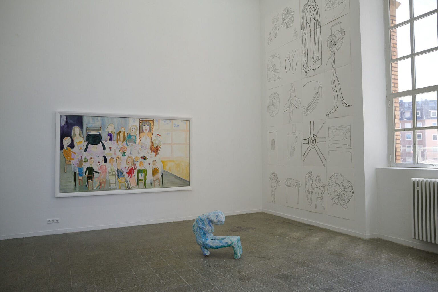 Foto: Ein Raum mit weißen Wänden, grauem, gefließten Boden und einem Fenster. An den Wänden eine Vielzahl flacher Objekte in unterschiedlichen Formaten und auf unterschiedlicher Höhe. Im Raum steht eine Skulptur auf dem Boden.