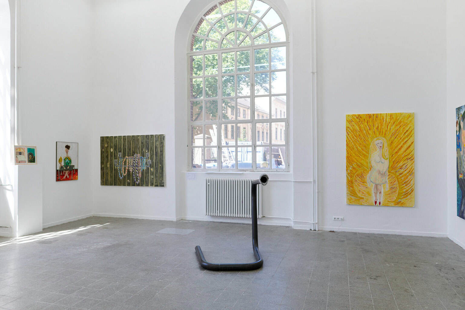 Foto: Ein Raum mit weißen Wänden, grauem, gefließten Boden und einem Fenster. An den Wänden eine Vielzahl flacher Objekte auf ähnlicher Höhe. Im Raum steht eine Skulptur auf dem Boden.