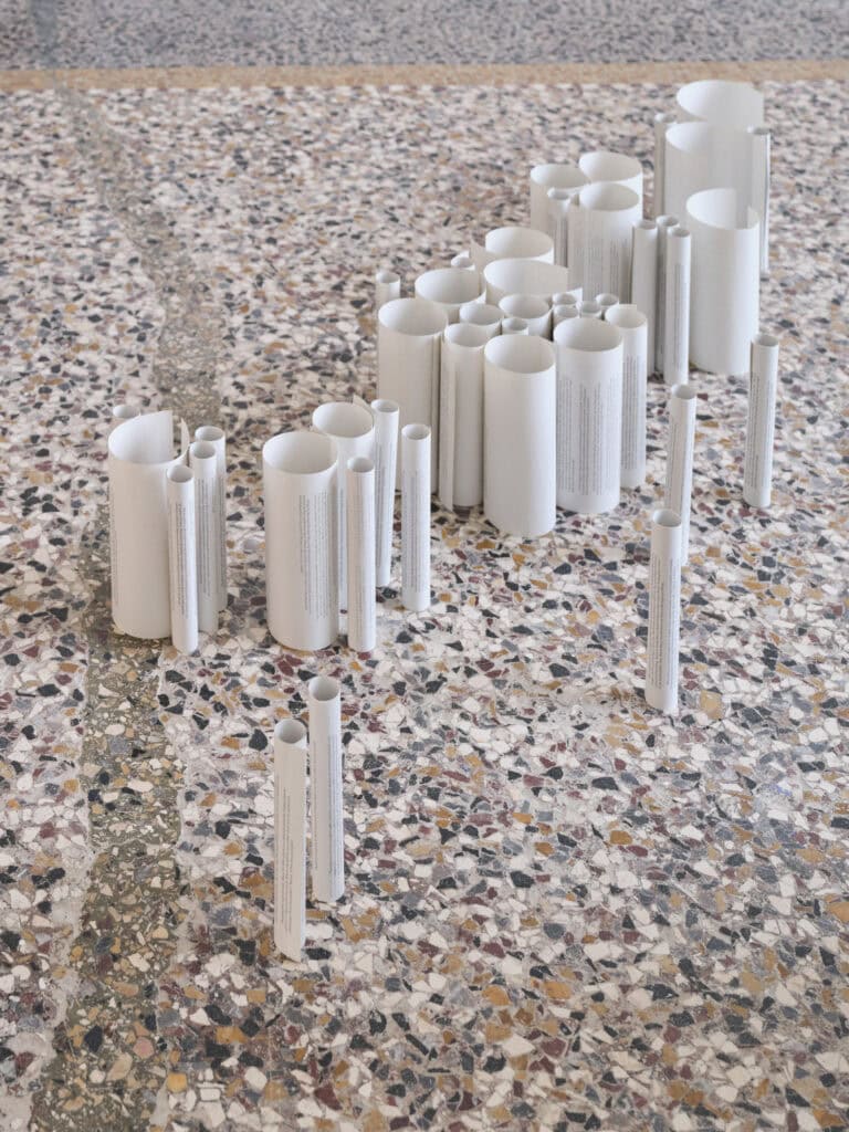 Foto: Nahaufnahme einer Kunstinstallation. Auf einem Terrazzoboden sind zusammengerollte bedruckte Papiere aufrecht aufgestellt
