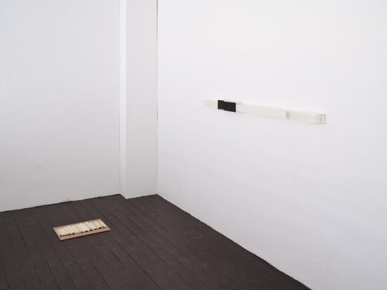 Foto: Aufnahme einer Ecke in einem Ausstellungsraum. Auf dem dunklen Holzboden liegt ein Brett mit einer wellenförmigen Arbeit. An der Wand ist ein längliches Element angebracht.