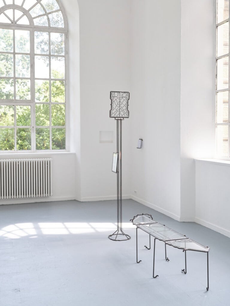 Foto: Aufnahme eines hellen Ausstellungsraums mit Rundbogenfenster. Im Raum stehen zwei Glas-Schmiedearbeiten