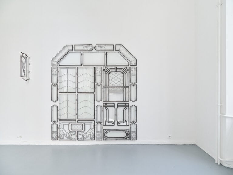 Foto: Aufnahme eines hellen Ausstellungsraums. An der Wand hängt eine großformatige flächige Glas-Schmiedearbeit