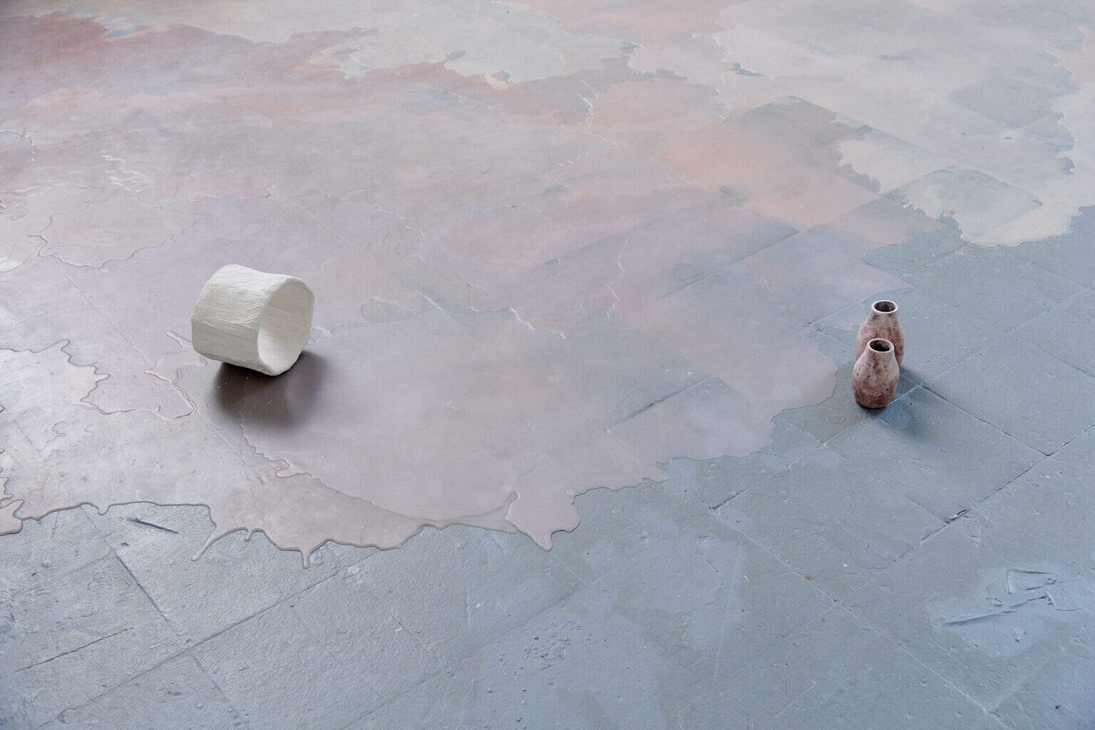 Foto: Detailaufnahme von drei Objekten auf grauem Boden. Teile des Bodens sind mit einer matten, rosafarbenen Masse bedeckt, die unebene, pfützenartige Konturen hat.