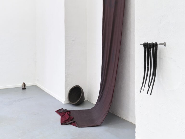 Foto: Detailaufnahme eines Kunstwerks. Langes dunkles Textilstück ist an der Wand befestigt und hängt bis zum Boden. Weitere schwarze abstrakte Kunstwerke sind auf dem Boden gestellt und an der Wand befestigt.