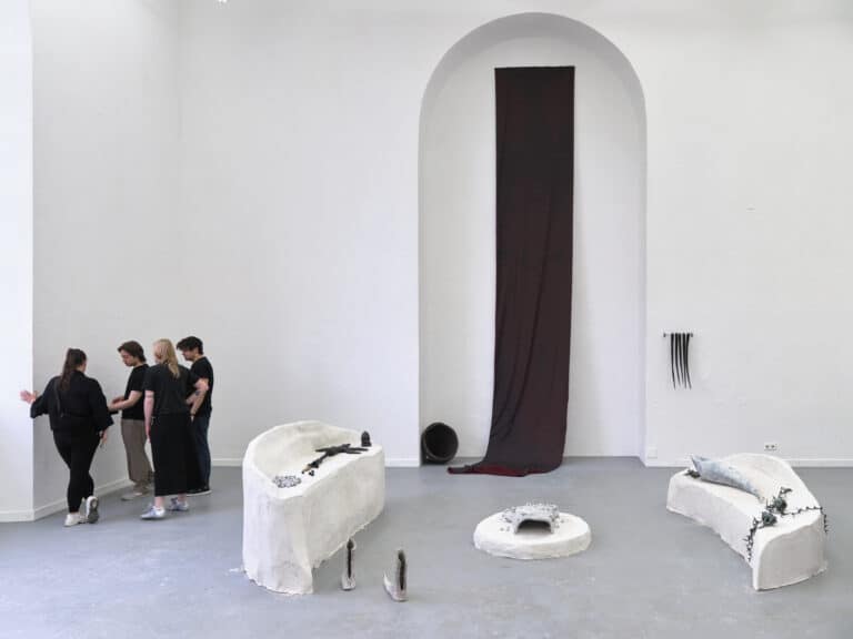 Foto: Aufnahme eines Ausstellungsraums mit weißen Wänden und grauem Boden. Mehrteilige Kunstinstallation hängt in einer bogenförmigen Wandnische und steht auf dem Boden davor. Langes schwarzes Textilstück ist an der Wand befestigt und hängt bis zum Boden. Drei organisch geformte Podeste stehen davor mit mehrteiligen Elementen.  Vier Personen stehen am linken Bildrand im Gespräch.