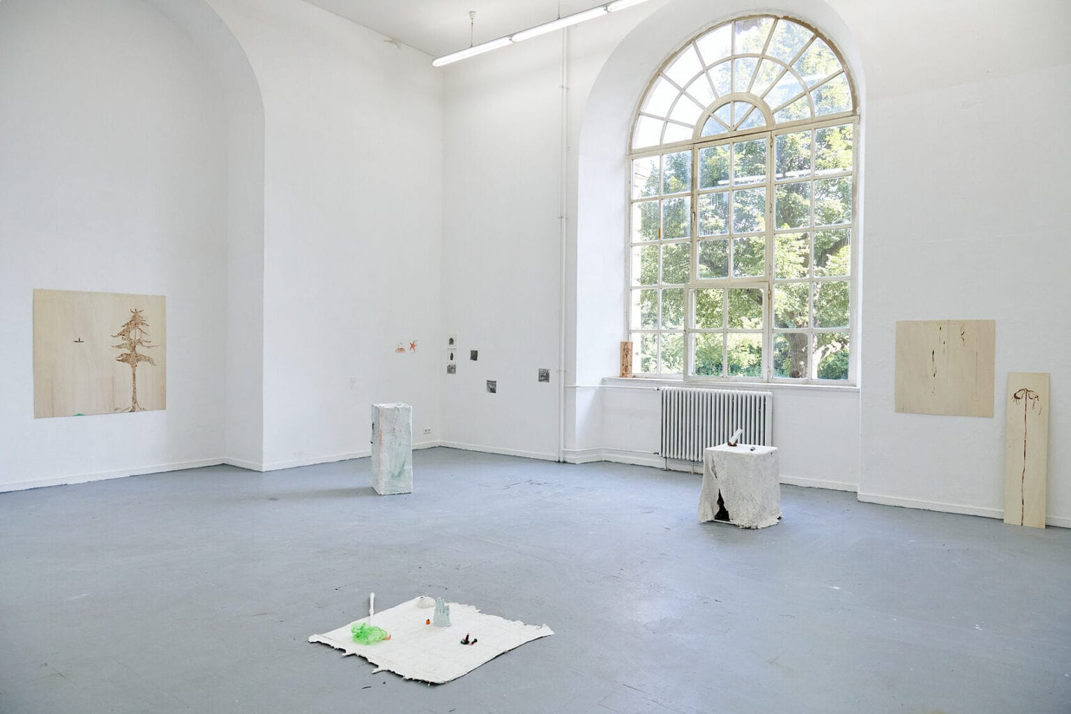 Foto: Ein Raum mit weißen Wände, grauem Boden und einem großen Fenster. Im Raum verteilt stehen mehrere Objekte. An der Wand hängen und lehnen mehrere Flache Objekte, alleine und in Gruppen.