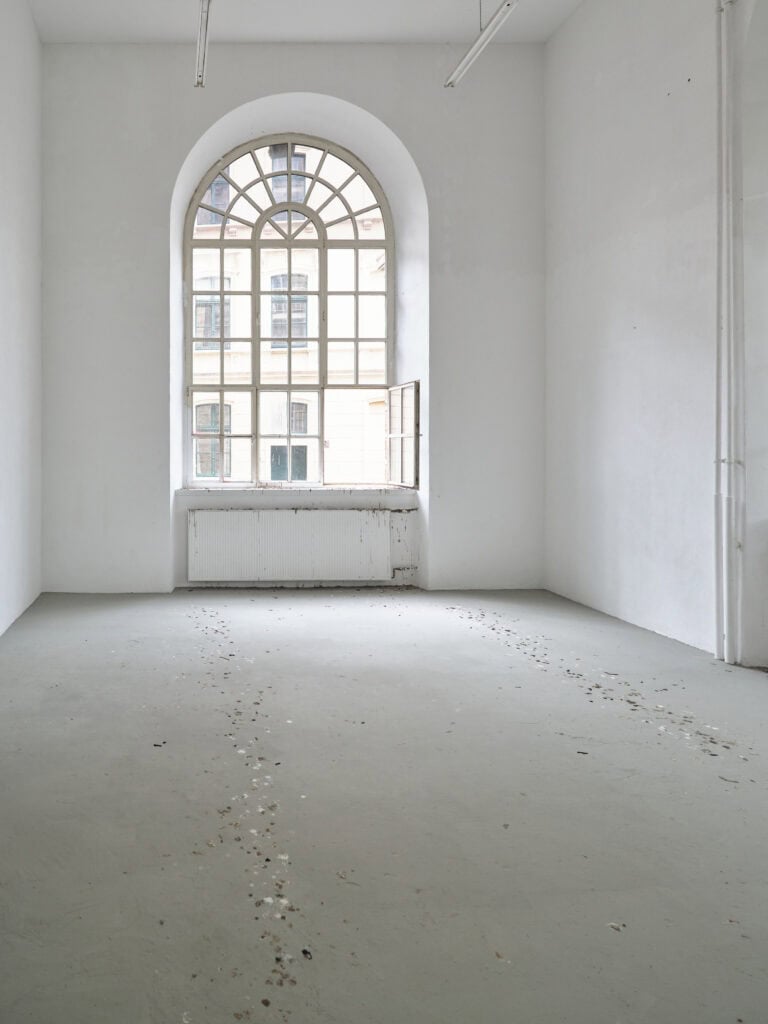 Foto: Ausstellungsraum mit großem Rundbogenfenster, weißen Wänden und grauem Boden. Ein Fensterflügel steht offen. Auf dem Boden, auf dem Heizkörper und der Fensterbank sind Flecken im Raum verteilt. Es sieht auf wie Vogeldreck.