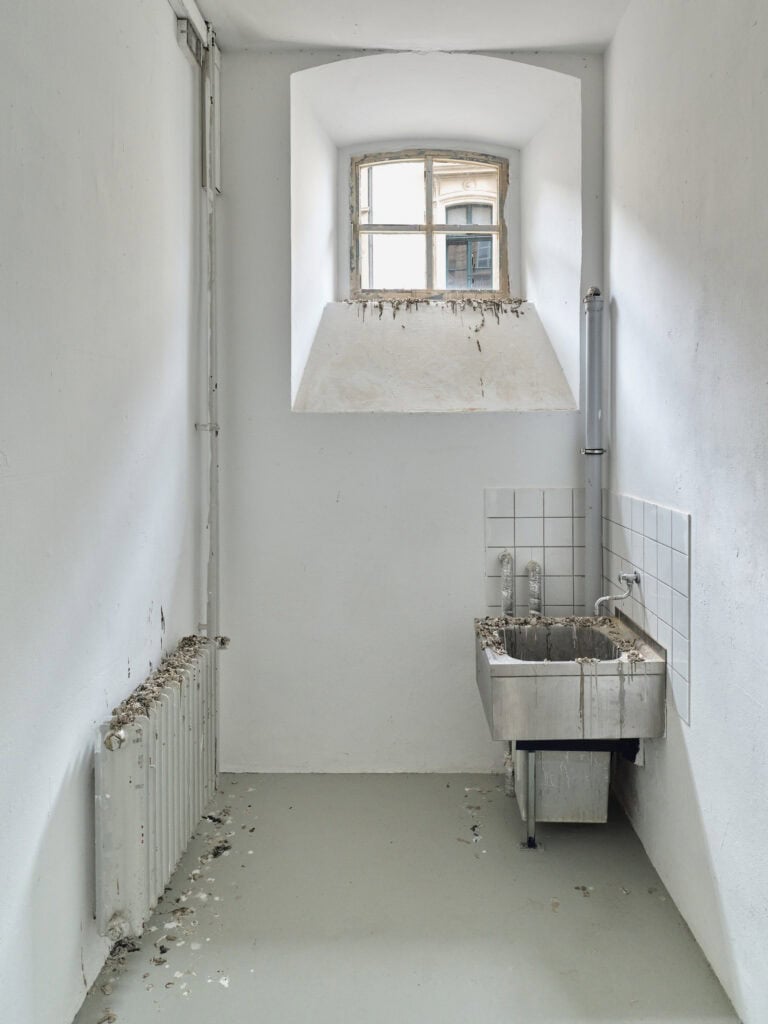 Foto: Ausstellungsraum mit kleinem Fenster, weißen Wänden und grauem Boden. In der rechten Ecke befindet sich ein Metallwaschbecken mit einem weißen Fliesenspiegel. Auf der rechten Seite hängt ein Heizkörper. Auf dem Boden, auf dem Heizkörper, der Fensterbank und auf dem Waschbecken sind Flecken im Raum verteilt. Es sieht auf wie Vogeldreck.