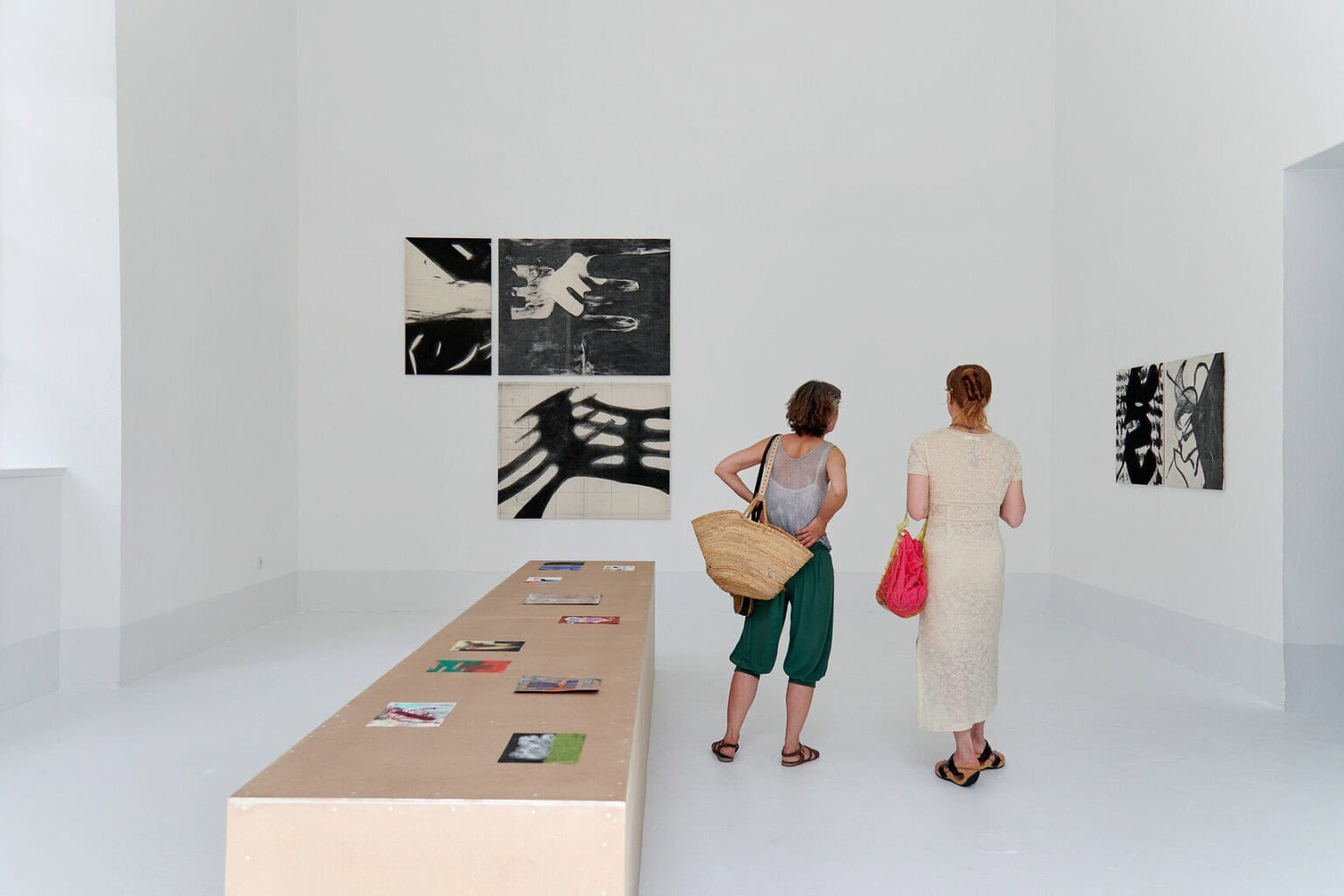 Foto: Ein Raum mit weißen Wänden, hellgrauem Boden und einem großen Fenster. Auf dem Boden steht ein länglicher, brauner Sockel auf dem kleinformatige Papierarbeiten liegen. An einer der Wände hängen Gruppen von Leinwandarbeiten im Mittelformat. im Raum stehen zwei Menschen.