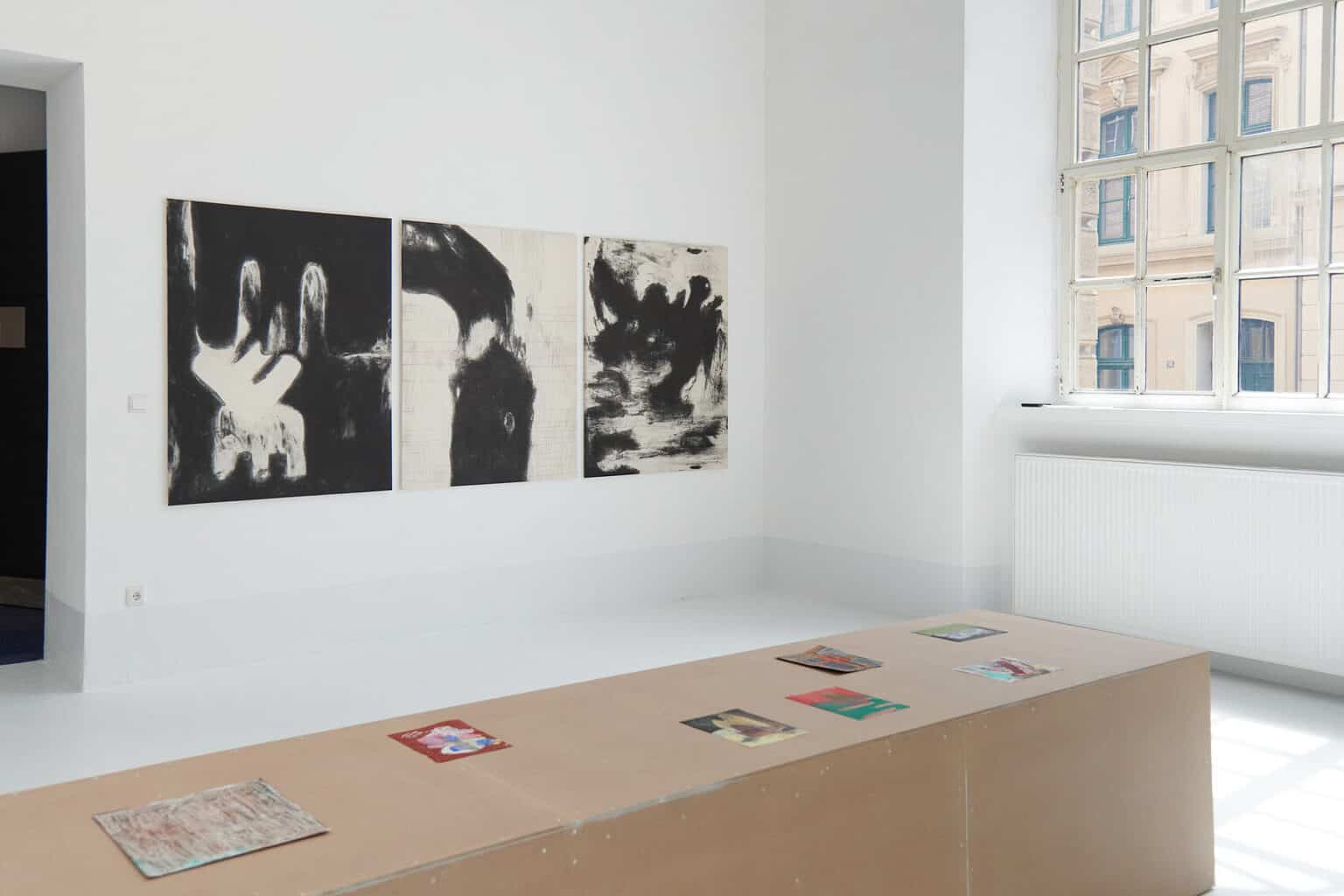Foto: Ein Raum mit weißen Wänden, hellgrauem Boden und einem großen Fenster. Auf dem Boden steht ein länglicher, brauner Sockel auf dem kleinformatige Papierarbeiten liegen. An einer der Wände hängen drei Leinwandarbeiten im gleichen Format, in einer Linie, nebeneinander.