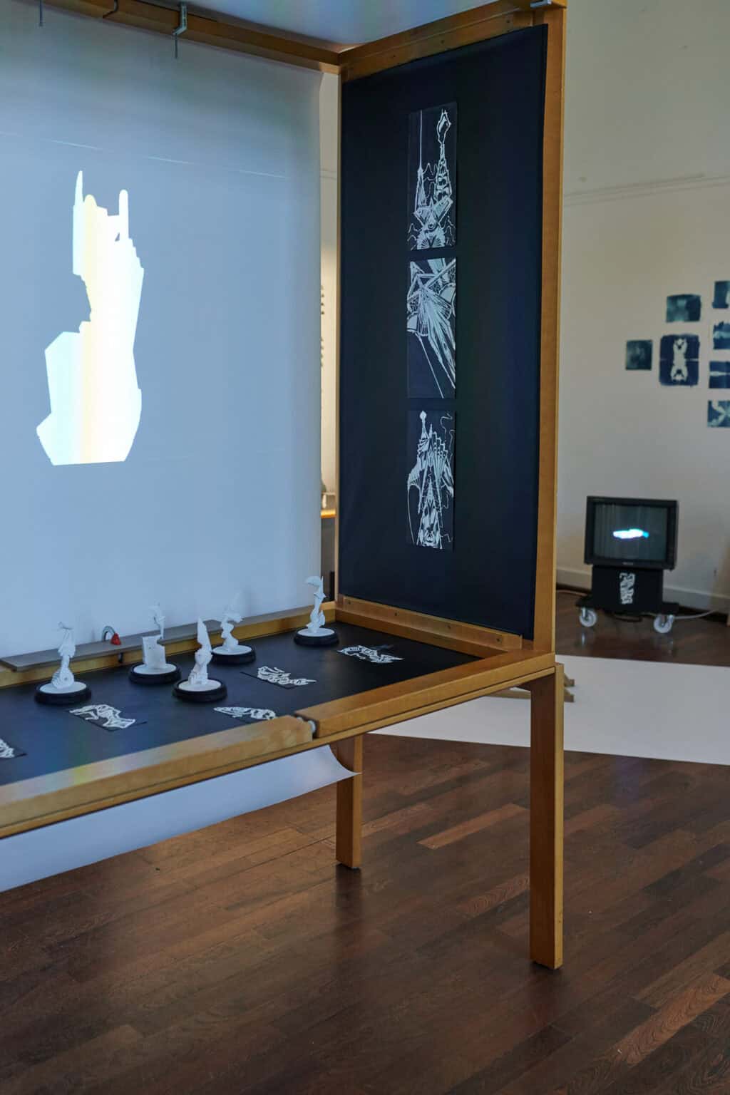 Foto: Ein Raum mit weißen Wänden und braunem Dielenboden. An den Wänden hängen und lehnen mehrere Gruppen von Objekten. Teile des Bodens sind in einer diagonalen Linie mit weißen Materialien bedeckt. Im Raum befinden sich mehrere Tische, Schaukästen und Konstruktionen, auf denen sich Objekte, Zettel und Technik befinden.