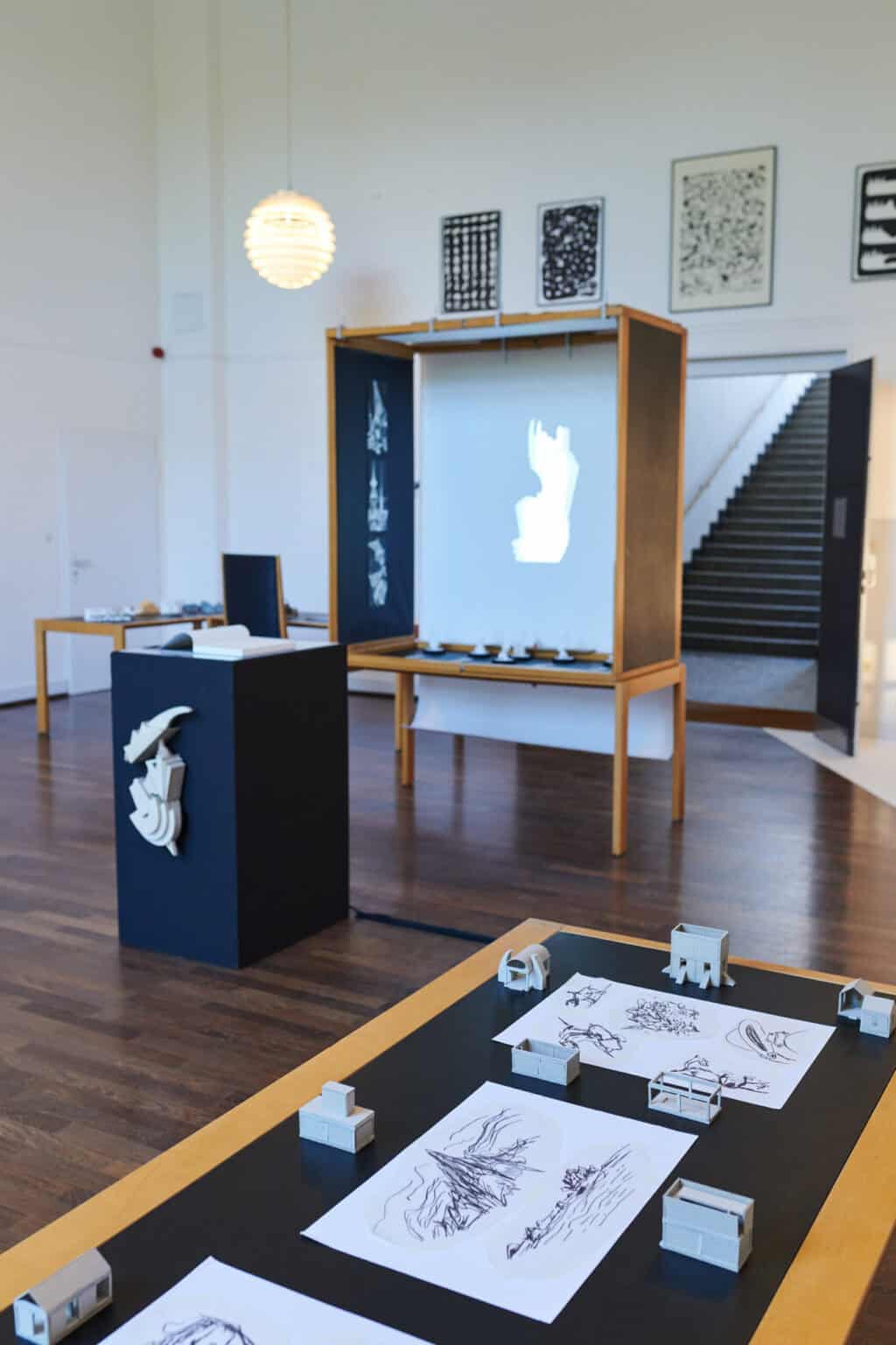 Foto: Ein Raum mit weißen Wänden und braunem Dielenboden. An den Wänden hängen und lehnen mehrere Gruppen von Objekten. Im Raum befinden sich mehrere Tische, Schaukästen und Konstruktionen, auf denen sich Objekte, Zettel und Technik befinden.