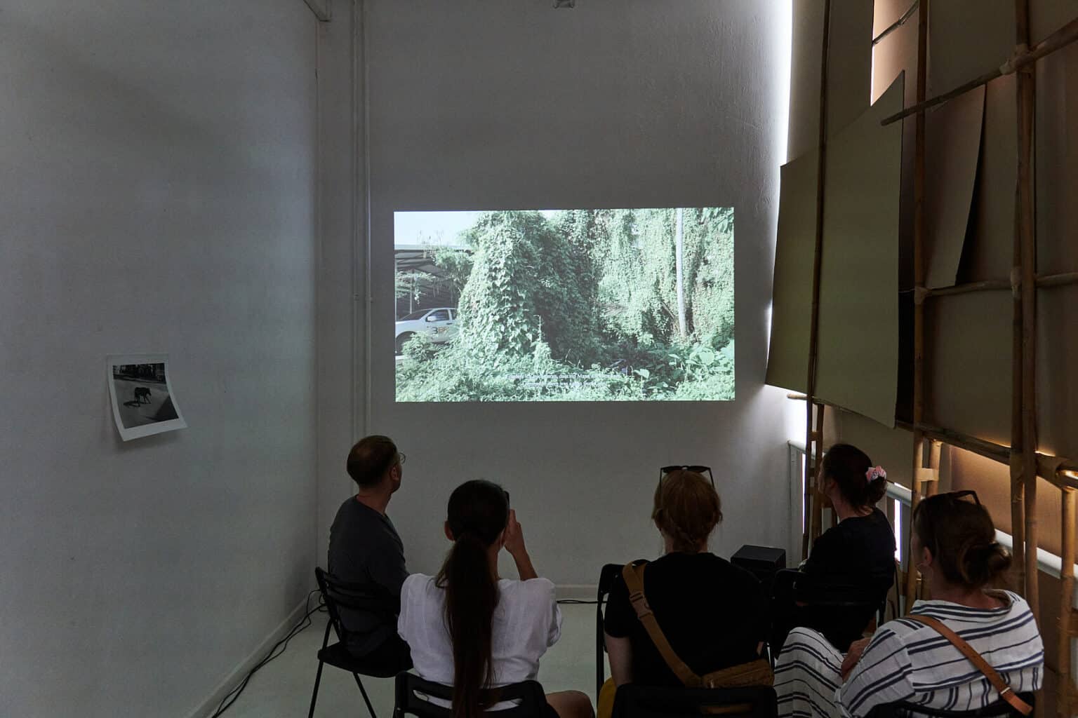 Foto: Ein abgedunkelter, schmaler Raum, in dem Menschen auf stuhlen sitzen. An der Wand ein bedrucktes Papier und eine Projektion. Durch eine der Wände, die aus ineinander gesteckten Pappen und Hölzern besteht, bricht etwas Licht.