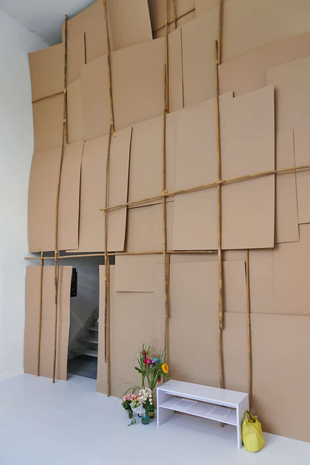Foto: Ein Raum mit weißen Wänden und grauem Boden. In den Raum ist eine Wand ein braunen Pappen eingezogen, die auf und an einer Konstruktion auf Stücken lehnen. Vor der Wand steht eine Bank oder ein Regal auf dem Boden, in dem Zettel liegen. Links daneben stehen Blumen.